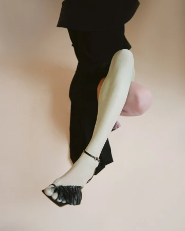 Close-up portrait of a model wearing black strappy heels and dark clothing against a pale seamless background styled by Tess Yopp.