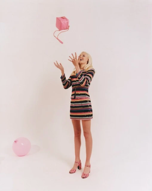 An adult woman with blonde hair is indoors wearing a striped dress and heels, tossing a pink purse in the air with a pink balloon.