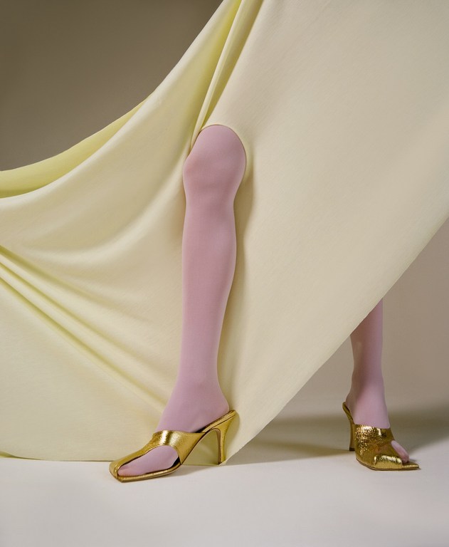 Abstract fashion shot of legs with pastel tights, gold high heels, and pale yellow fabric in a photo studio setting. Neutral colors.