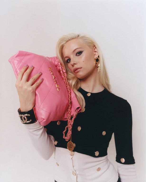 A woman with platinum blonde hair holds a pink quilted Chanel bag with gold chain in a studio portrait against a white background.