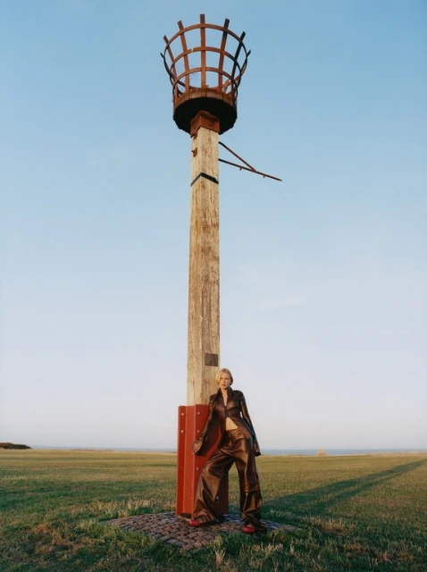 Woman in brown suit poses next to vintage wooden pylon on grassy field under blue sky in editorial fashion shot. 