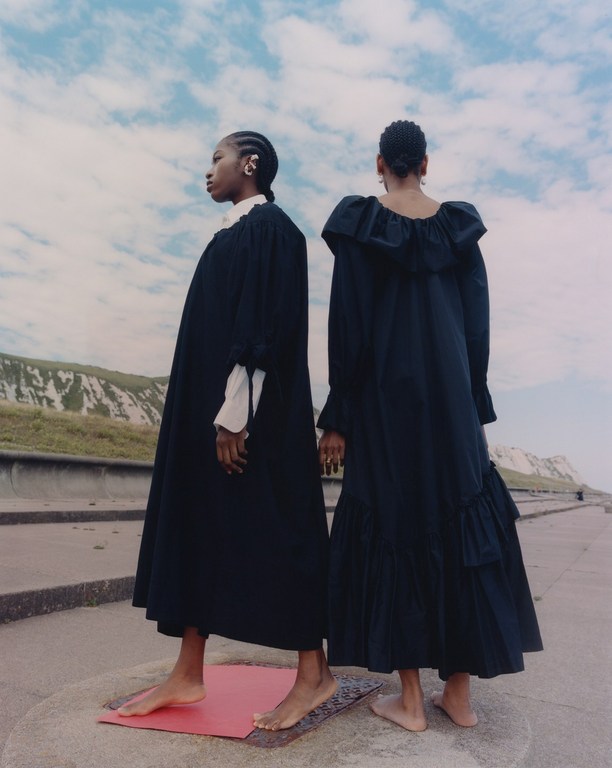 Two Black women stand on a platform in long black dresses with a coastal backdrop under a cloudy sky, fashion-editorial style.