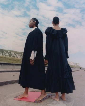 Two Black women stand on a platform in long black dresses with a coastal backdrop under a cloudy sky, fashion-editorial style.