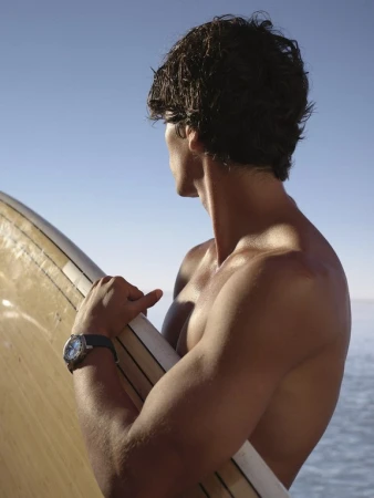 Shirtless man with dark hair holding a surfboard outdoors against a clear blue sky, styled by Tess Yopp in London.