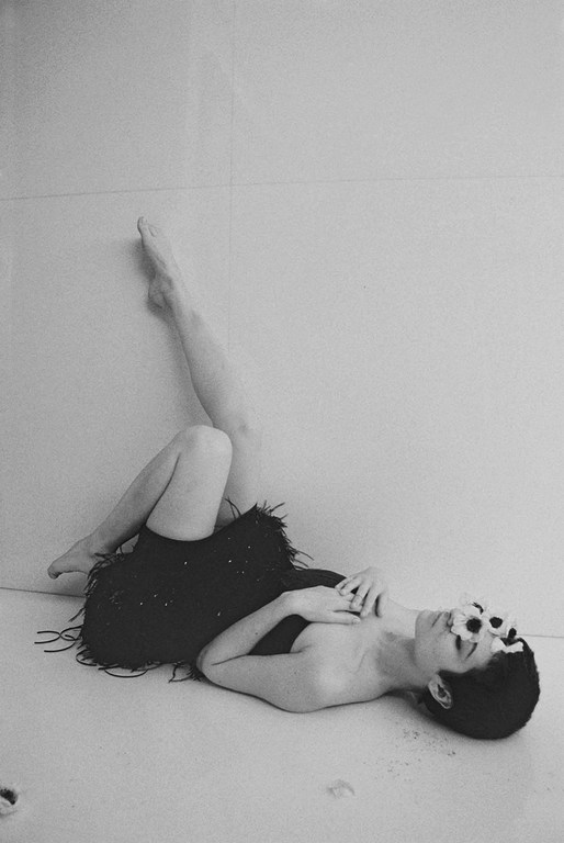 Monochrome portrait of a woman in a feather dress lying on the floor in a white-walled studio, gazing upward with a serene expression.