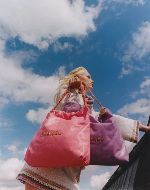 Fashion shoot featuring a model wearing a light-colored coat and holding pink and purple Chanel bags against a bright sky backdrop.