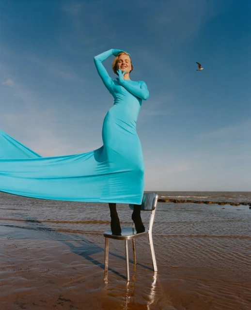 A woman in a blue full-body suit poses on a chair at the beach against a blue sky, a long flowing fabric detail and a gull in flight.