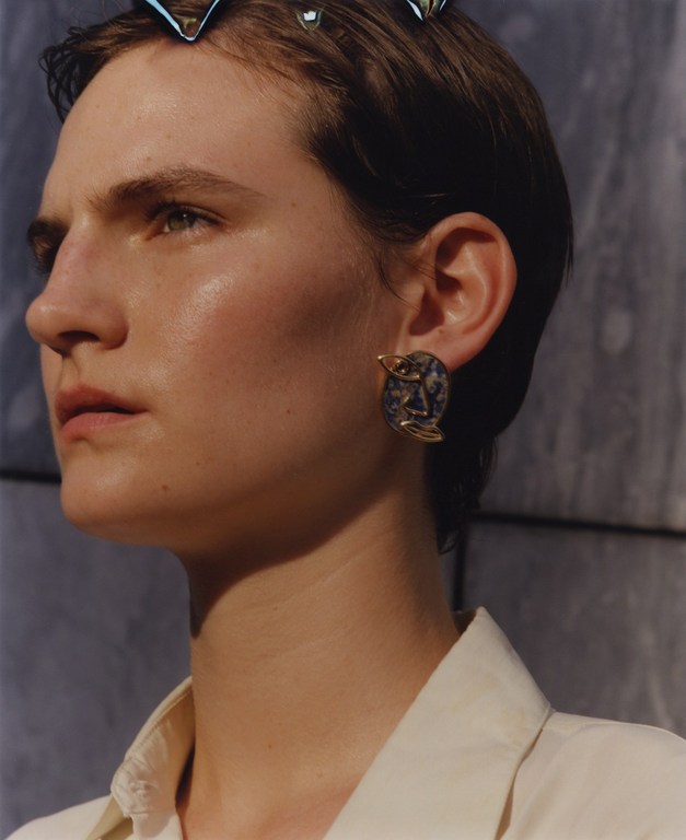 Close-up of a young woman with short hair, wearing a statement earring and an off-white collared shirt against a textured gray backdrop.