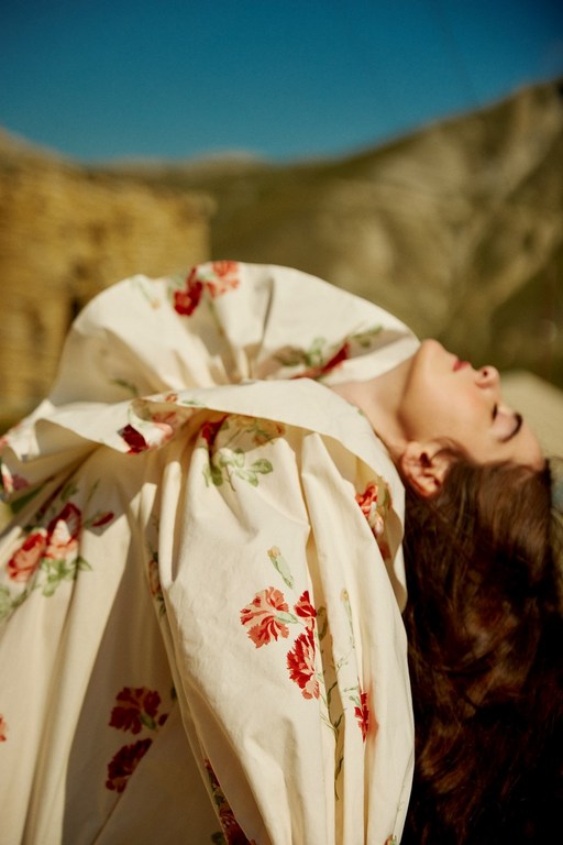 Woman in a floral print dress reclines outdoors against a hillside under a clear blue sky in a fashion-style photograph.