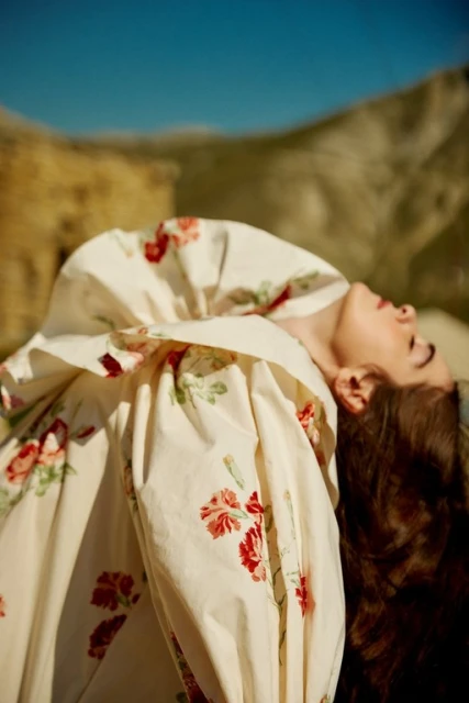 Woman in a floral print dress reclines outdoors against a hillside under a clear blue sky in a fashion-style photograph.