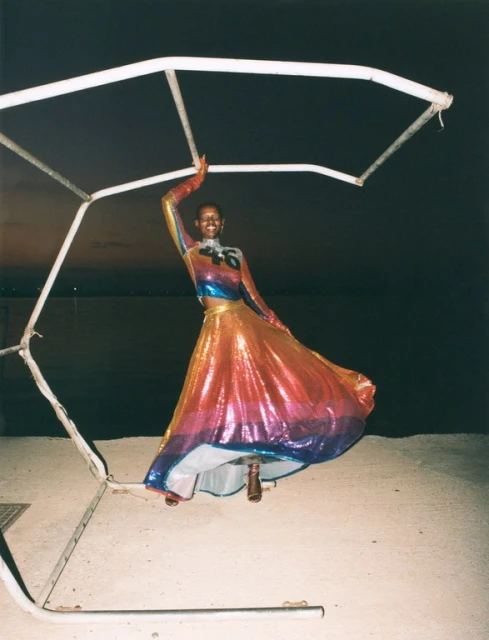 Fashionable woman in iridescent skirt and top strikes a pose next to metal framework on concrete in this full shot against a night sky.