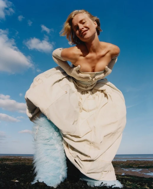 Woman poses in an off-the-shoulder light-beige garment with light-beige gloves, a blue, fur-like skirt, against a blue sky with clouds.