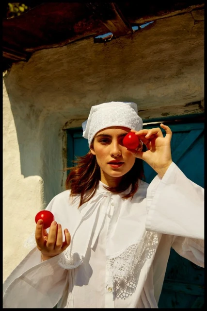 A woman in a white cap and shirt holds red tomatoes, one covering her eye, against a whitewashed wall with a blue door.