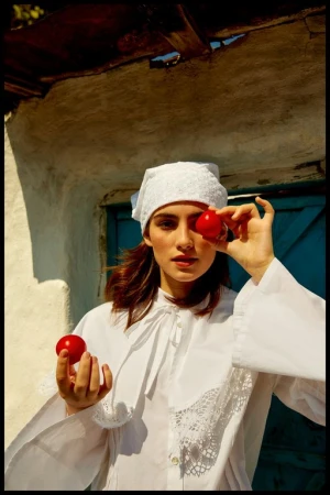 A woman in a white cap and shirt holds red tomatoes, one covering her eye, against a whitewashed wall with a blue door.