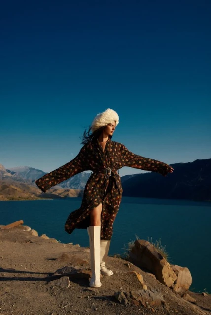Woman poses outdoors in patterned coat and tall white boots, set against mountains and a blue lake under a clear blue sky. Fashion.