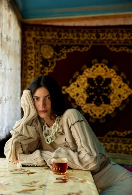 Brunette woman poses indoors wearing cream knitwear, a statement necklace, sitting at a table with a glass of tea and patterned textile.
