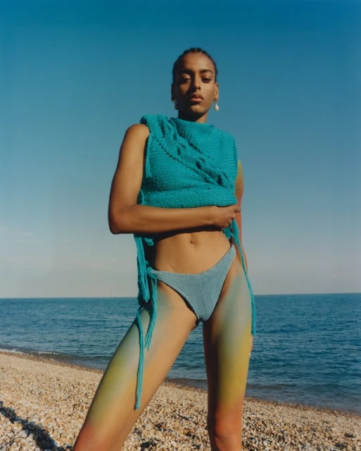 Woman on a rocky beach wearing a teal knit top and bikini bottom, with blue sky and water visible in the background on a sunny day.