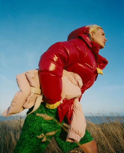 Outdoor fashion shot of a blonde adult female wearing a red puffer jacket and green shorts, holding a beige jacket against a blue sky.