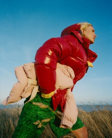 Outdoor fashion shot of a blonde adult female wearing a red puffer jacket and green shorts, holding a beige jacket against a blue sky.