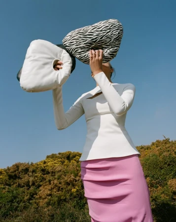 Model wearing a white top and pink skirt holding two abstract pillows against a clear blue sky and foliage background