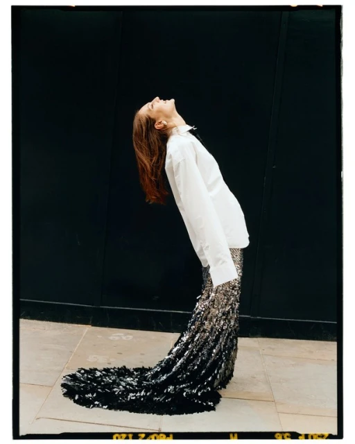 Woman in stylish outfit, white shirt and sequin skirt, posing with head tilted back against a dark wall, fashion editorial shot.