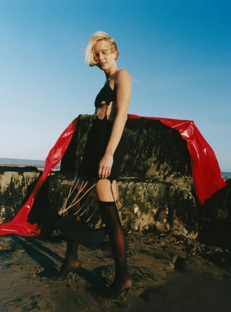 Blonde woman in black dress and stockings stands on a rocky beach with blue sky and red fabric accent, fashion editorial outdoor shot.