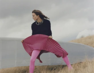 Model wearing a black sweater and bright pink pleated skirt holding out fabric against an overcast sky and grassy hill