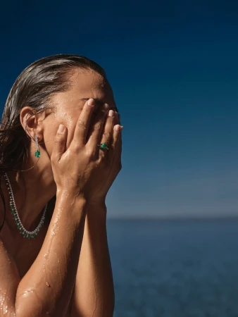 Wet-haired woman covering her face with both hands against a deep blue sea background, wearing emerald jewelry.