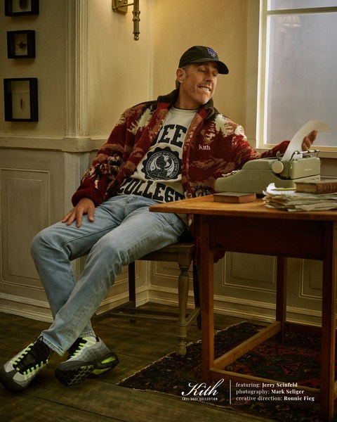 Jerry Seinfeld sits at a desk with a typewriter in a magazine editorial shot, wearing a college sweatshirt, denim jeans and sneakers.