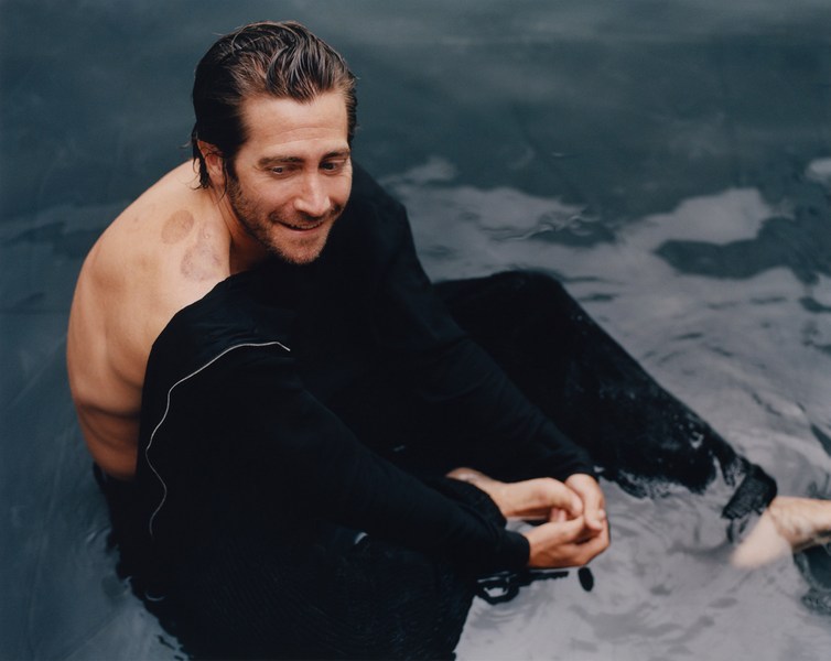 Jake Gyllenhaal sits in water with black pants and no shirt, smiling softly, showcasing his relaxed pose and natural setting in the water.