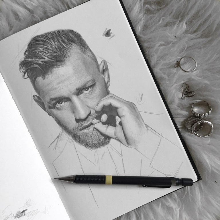 Pencil drawing of fair-skinned adult male with beard, sketched in profile on notebook paper, with pencil and rings on furry surface.