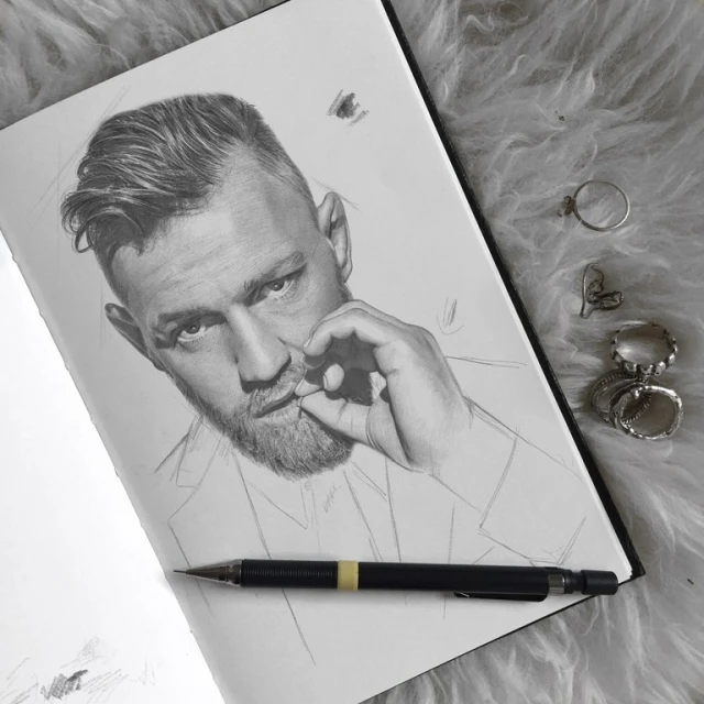 Pencil drawing of fair-skinned adult male with beard, sketched in profile on notebook paper, with pencil and rings on furry surface.