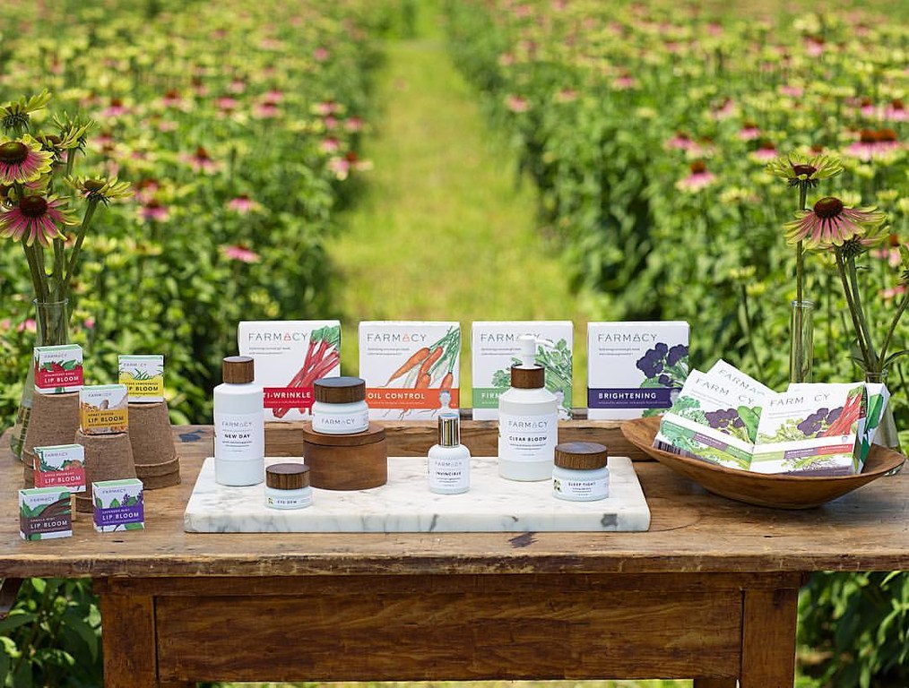 Farmacy beauty product line arranged on a wooden table in a field of purple coneflowers, featuring serums, creams, and lip bloom products.