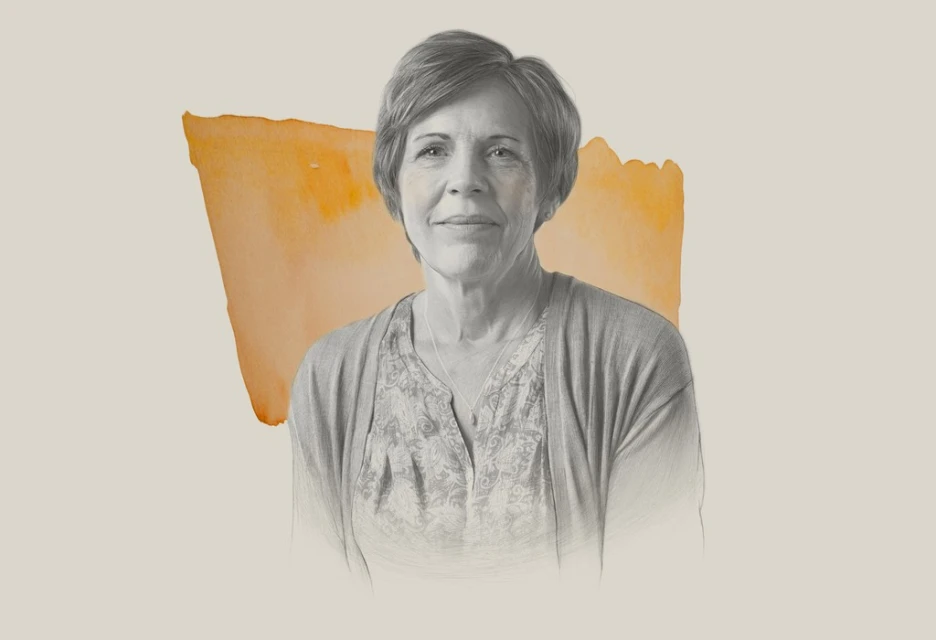 Portrait of a mature woman with short grey hair wearing a patterned blouse and cardigan against a painted beige backdrop.