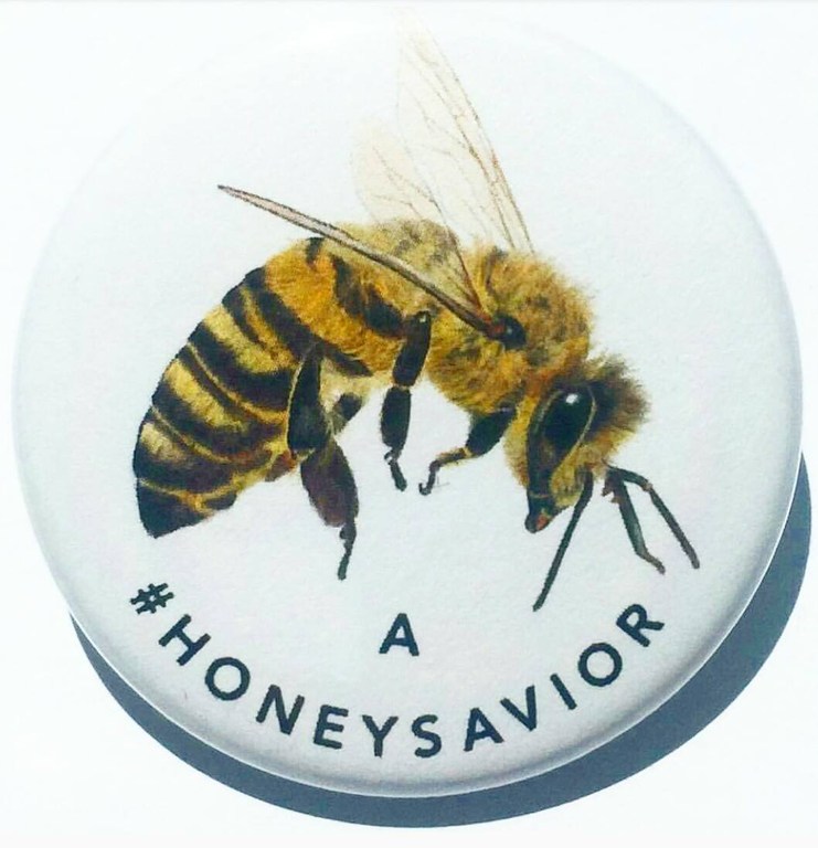 Honey bee on a white button with the text '#HONEYSAVIOR'. Close-up of the insect against a clean, minimalist background.