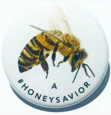 Honey bee on a white button with the text '#HONEYSAVIOR'. Close-up of the insect against a clean, minimalist background.