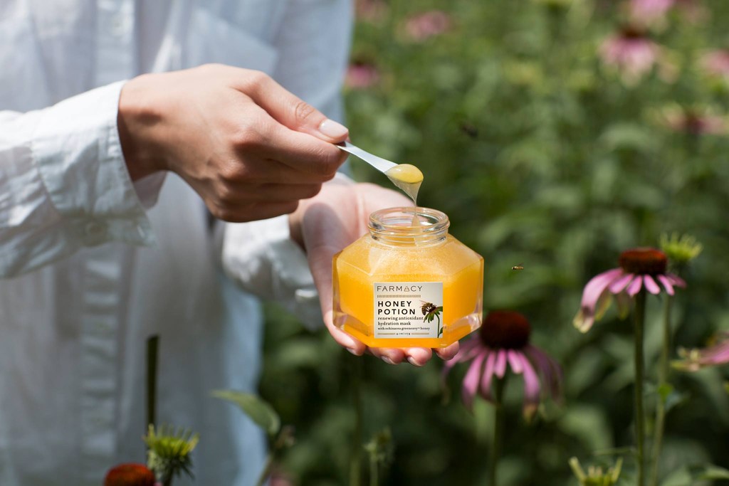 Close-up of hands holding honey-filled Farmacy Honey Potion jar with spoon, in a garden setting with purple coneflowers and blurred background.