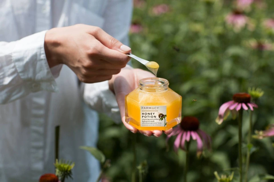 Close-up of hands holding honey-filled Farmacy Honey Potion jar with spoon, in a garden setting with purple coneflowers and blurred background.