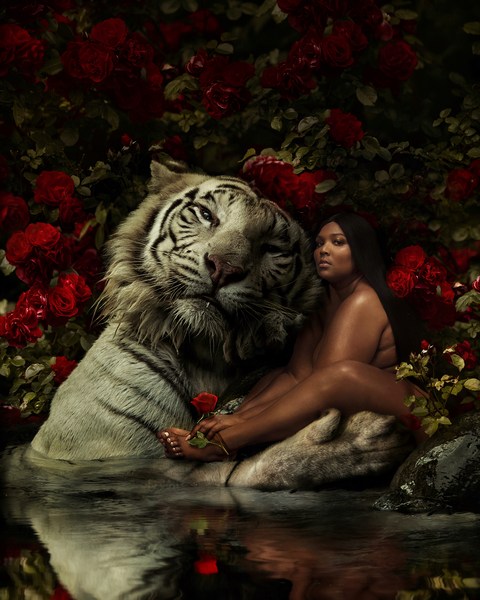 Adult woman with long dark hair seated next to a white tiger in a pool of water surrounded by red roses and greenery in soft lighting.