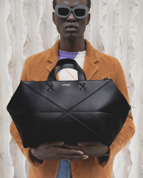 Dark-skinned male model in stylish sunglasses and an orange coat holds a black geometric Loewe bag against a textured wall.