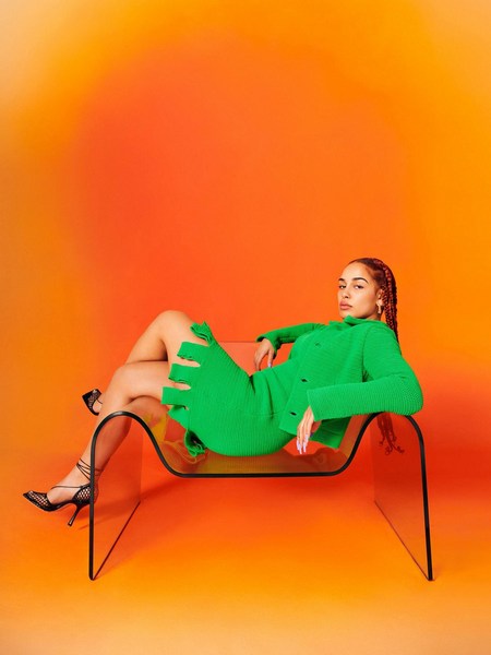 An adult female model with long hair lies in a clear plastic chair, wearing a green dress and black heels, against an orange studio backdrop.