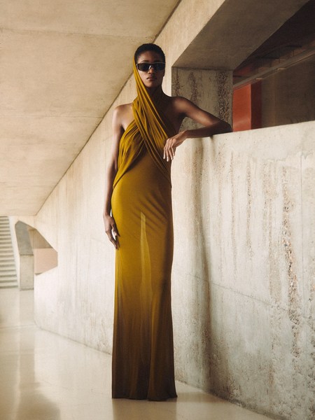 A Black woman stands in a mustard-colored gown with sunglasses in a concrete structure, bathed in natural light.