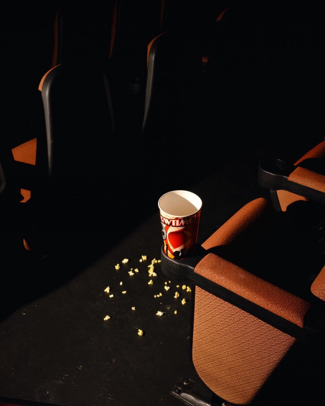 An image inside a dark movie theatre shows a cup of popcorn placed in a cup holder of a tan-coloured chair, with some popcorn spilled around.