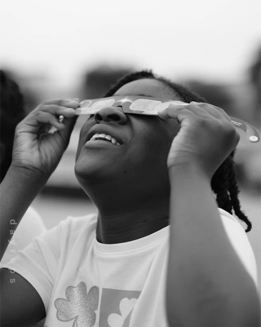 Monochrome portrait of a woman wearing eclipse glasses, looking up, with a faint sky and blurred background outside during daylight.