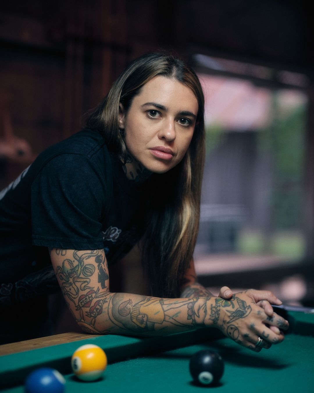 Portrait of a tattooed adult female leaning on a green billiard table with balls, inside a dimly lit, wood-paneled recreation room.