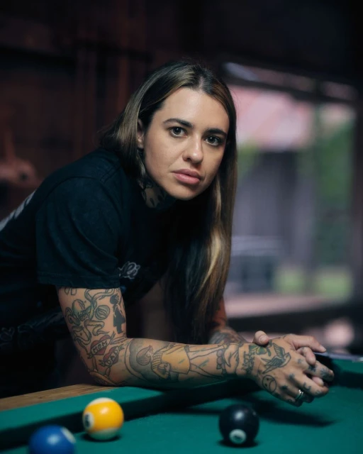 Portrait of a tattooed adult female leaning on a green billiard table with balls, inside a dimly lit, wood-paneled recreation room.