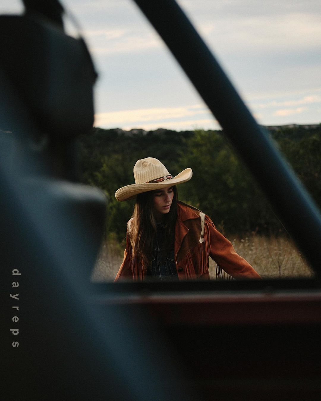 Woman in cowboy hat and fringed jacket poses in rural setting, seen through blurred vehicle frame, dayreps logo at left.