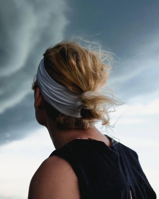 From behind, a person with blonde hair and a white headband gazes upward at dramatic storm clouds in an outdoor setting.