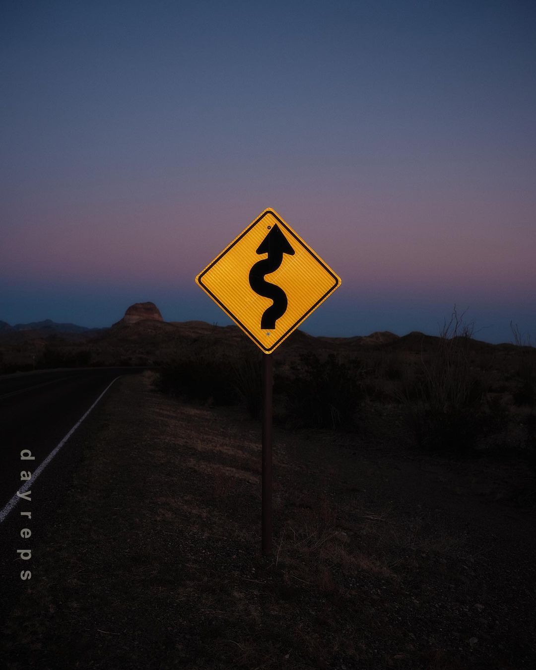 A yellow traffic sign warns of a winding road ahead at dusk, with a dark asphalt road and hilly landscape visible.