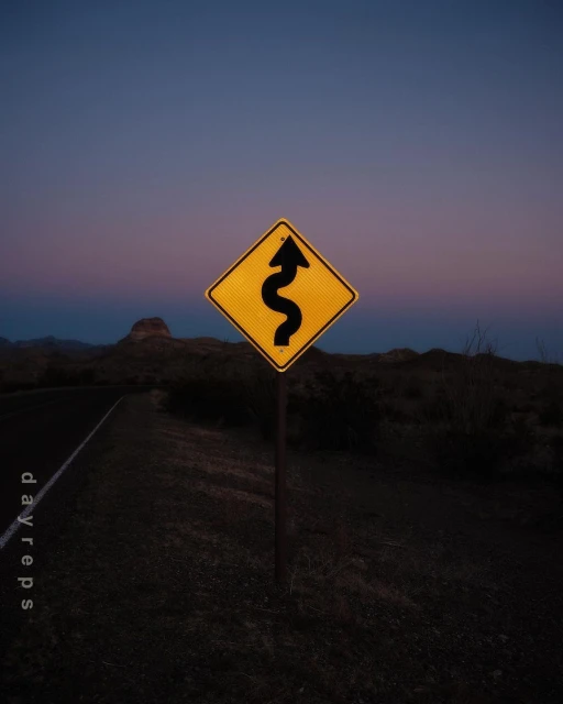 A yellow traffic sign warns of a winding road ahead at dusk, with a dark asphalt road and hilly landscape visible.